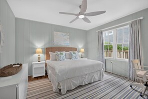 A bedroom with a bed, nightstands, a ceiling fan, a window with curtains, and a chair. - Baba's Beach House - Oceanfront Family Cottage (Jacksonville Beach)