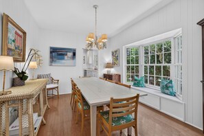A dining area with a long table, chairs, a chandelier, and a large window with a view of greenery. - Baba's Beach House - Oceanfront Family Cottage (Jacksonville Beach)