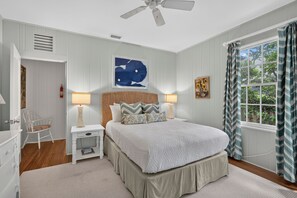 Un dormitorio con cama, lámparas de noche, ventilador de techo, ventana con cortinas, una obra de arte en la pared y una pequeña mesa con una silla.