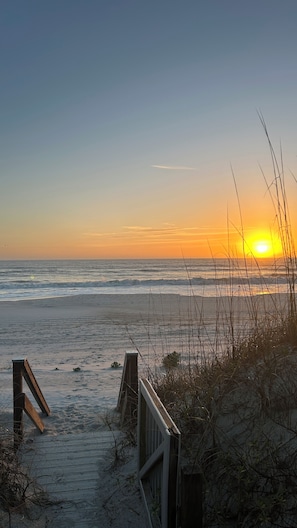 Une scène de plage avec une promenade en bois menant à l’eau, des vagues et un coucher de soleil.