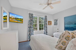 Une chambre à coucher avec un lit, un ventilateur de plafond, une fenêtre avec des rideaux, un tableau et un téléviseur fixé au mur.