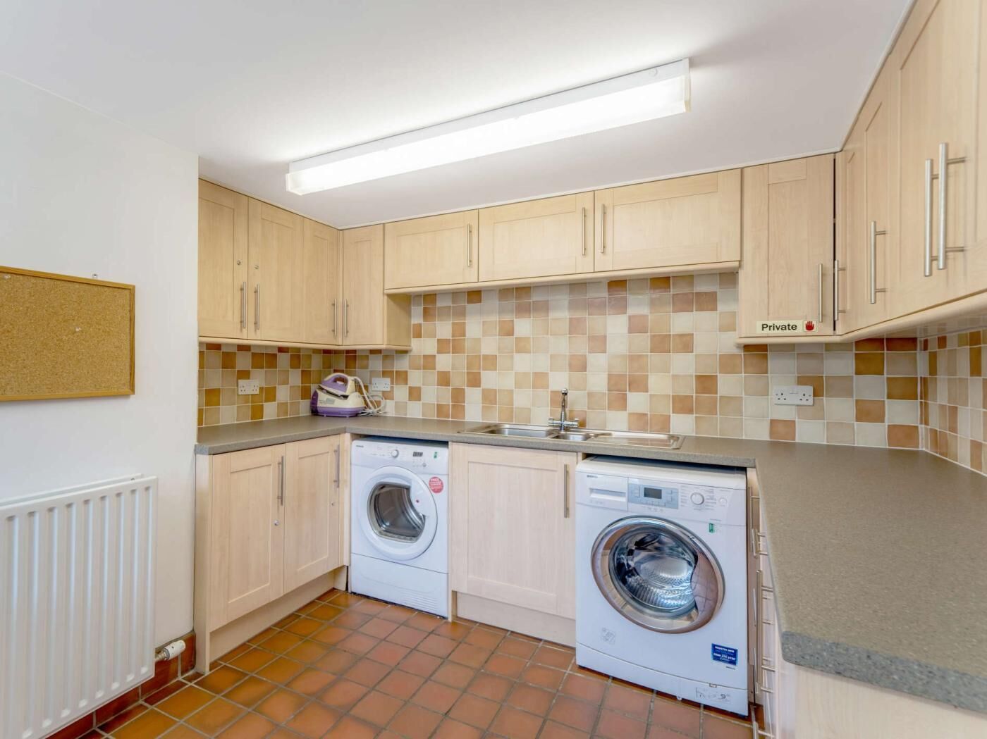 Cocina compacta con dos lavadoras, salpicadero de azulejos y armarios de madera clara.