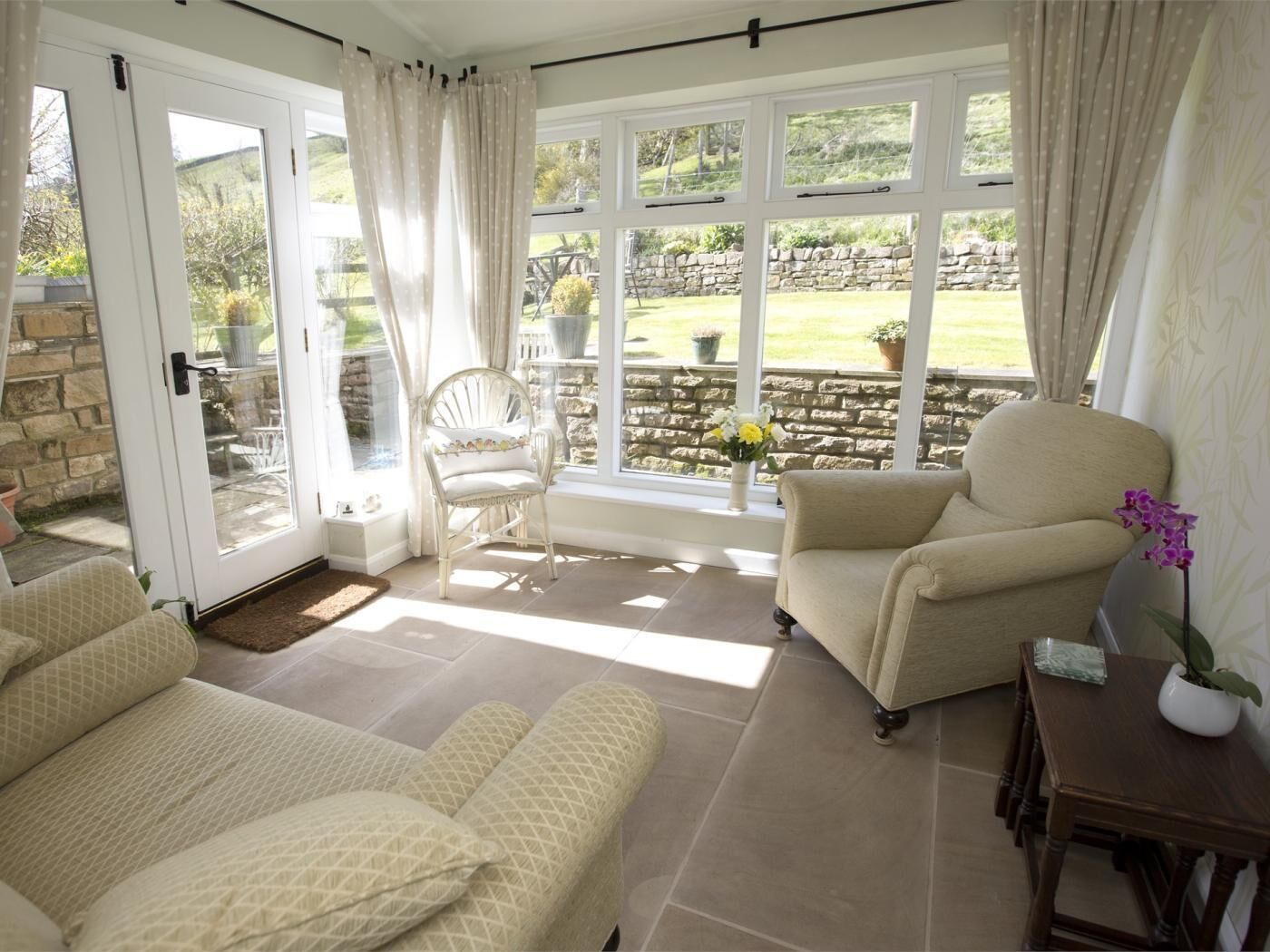 A room with a sofa, a chair, a table, and a view of a garden through large windows.