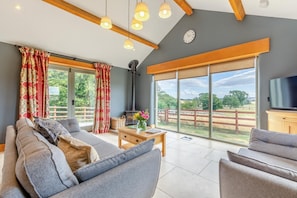 Ein geräumiges Wohnzimmer mit einer Couch, Sesseln, einem Couchtisch und einem Fernseher. Ein großes Fenster bietet Blick ins Freie.