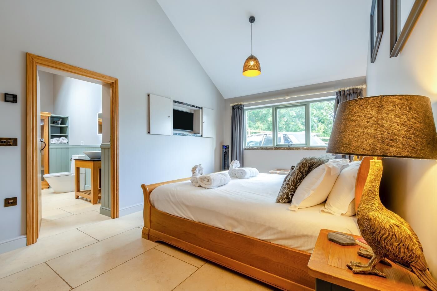 A hotel room with a wooden bed, a desk with a lamp, a television on the wall, and a window with a view of the outside.