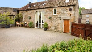 A stone building with a tiled roof, a wooden door, and a brick courtyard with a table and chairs. - 1 Bed in Ripley (oc-g0127) (Kirkby Malzeard)