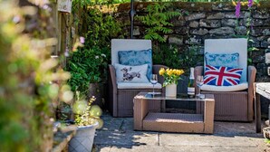 A patio with wicker furniture, two chairs with cushions, a table with a vase of flowers, and a stone wall. - 1 Bed in Pickering (oc-g0120) (Rosedale Abbey)