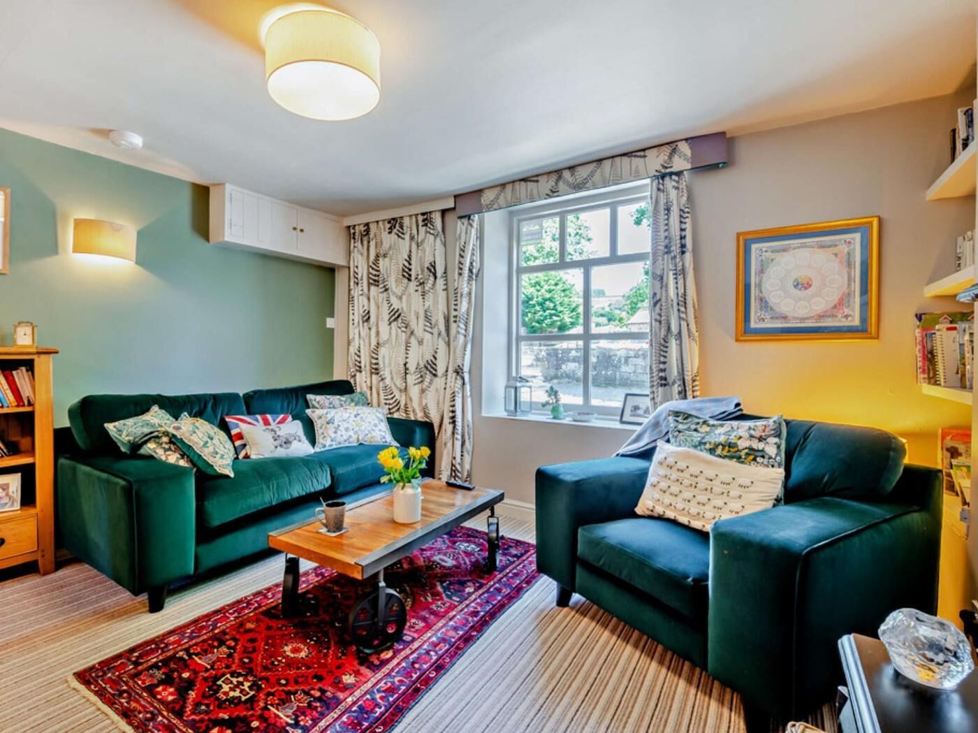 A living room with two dark green sofas, a wooden coffee table, a red patterned rug, a window with curtains, and a bookshelf.