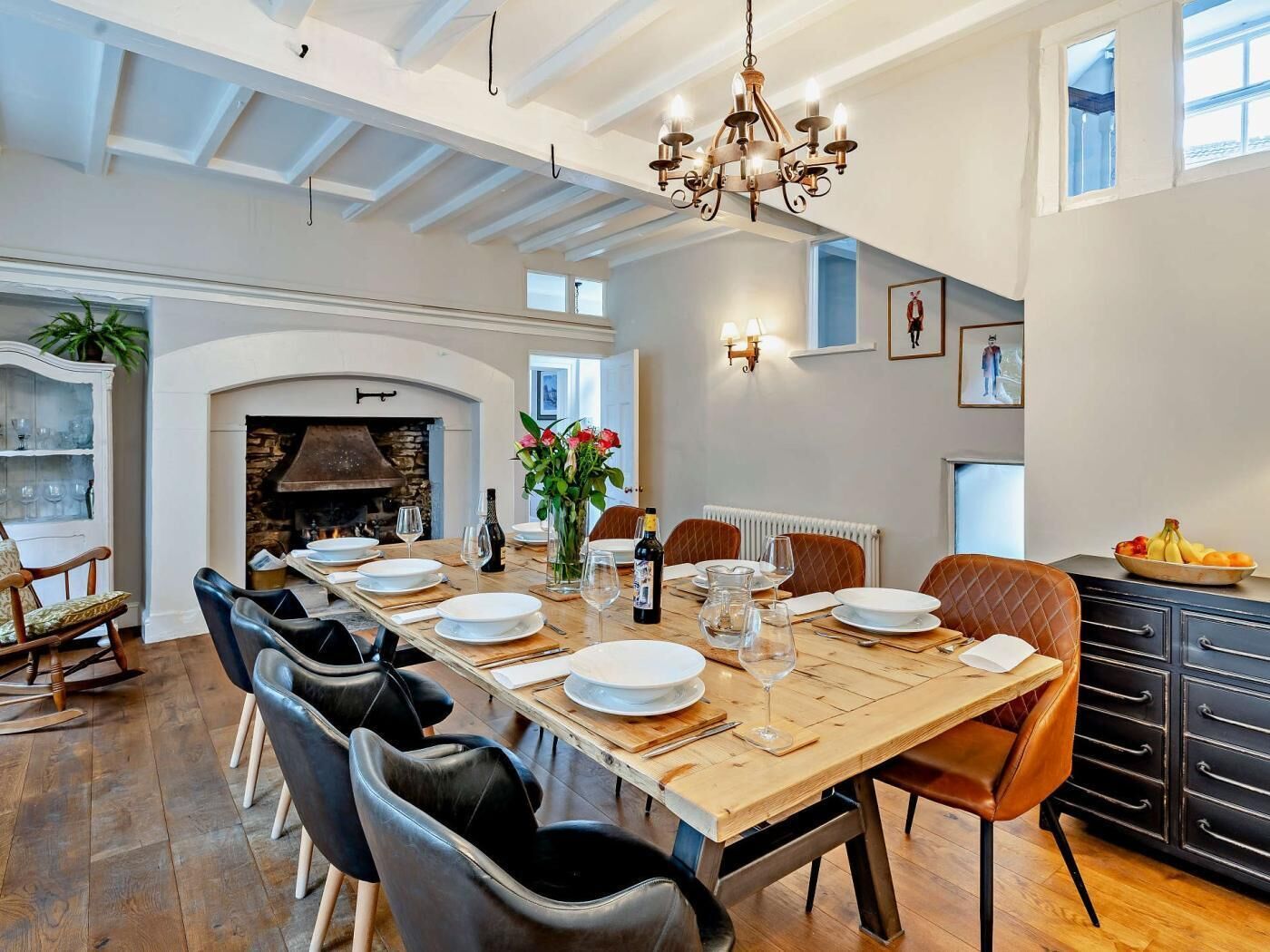 A dining room with a long table set for six, surrounded by chairs, a chandelier, and a fireplace.