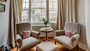 Een kamer met twee fauteuils, een klein rond tafeltje met een bloemenvaas en uitzicht op een stenen gebouw door grote ramen.