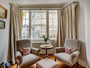 Ein Zimmer mit zwei Sesseln, einem kleinen runden Tisch mit Blumenvase und Blick auf ein Steingebäude durch große Fenster.