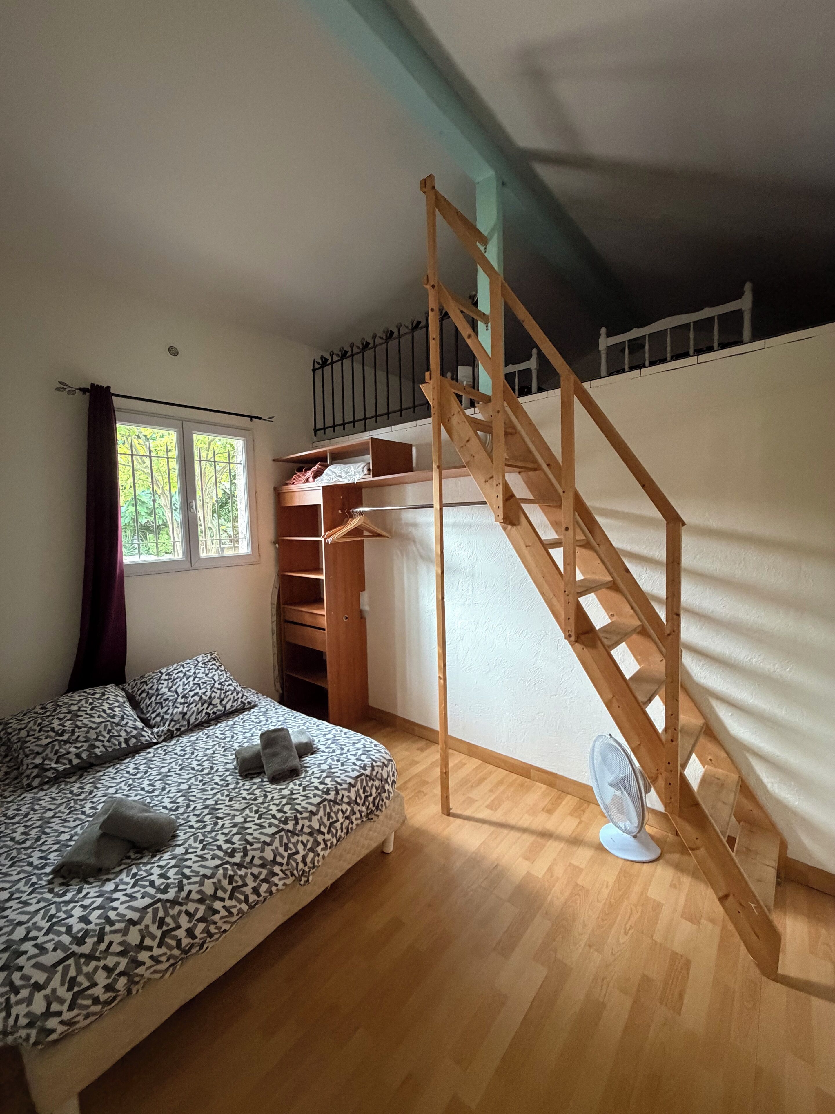 A room with a bed, a wooden staircase, a window, and a fan.