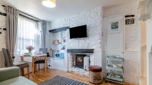 A cozy living room with a fireplace, a flat-screen TV mounted on a textured wall, a dining area with a table and chairs, and a kitchen area with a microwave. - 1 Bed in Brixham (oc-73940) (Brixham)