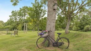Ein Fahrrad, das an einen Baum gelehnt ist, mit einem Blumenkorb, einer Holzbank und einem Vogelhäuschen in einer grünen Außenanlage.