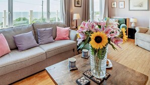 A living room with a sofa, a coffee table with a vase of flowers, and a view of the outdoors through large windows. - 4 Bed in Bridgend (oc-74236) (Ogmore-by-Sea)