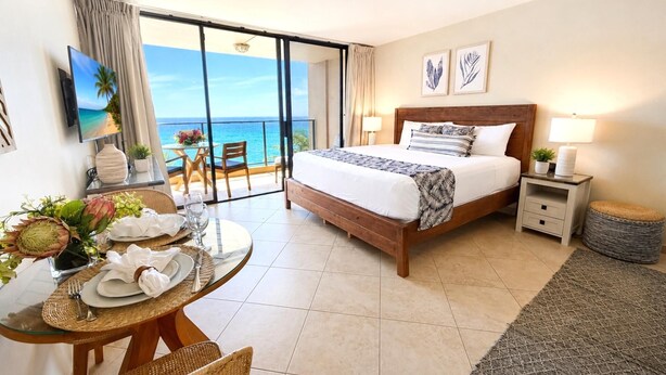Una camera d'albergo con un letto, un tavolo da pranzo con sedie, un balcone vista oceano e una TV montata a parete.