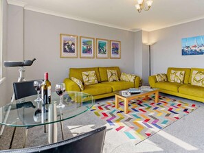 Ein Wohnzimmer mit einem gelben Sofa, einem Glastisch, einem Teleskop und gerahmten Kunstwerken an der Wand.
