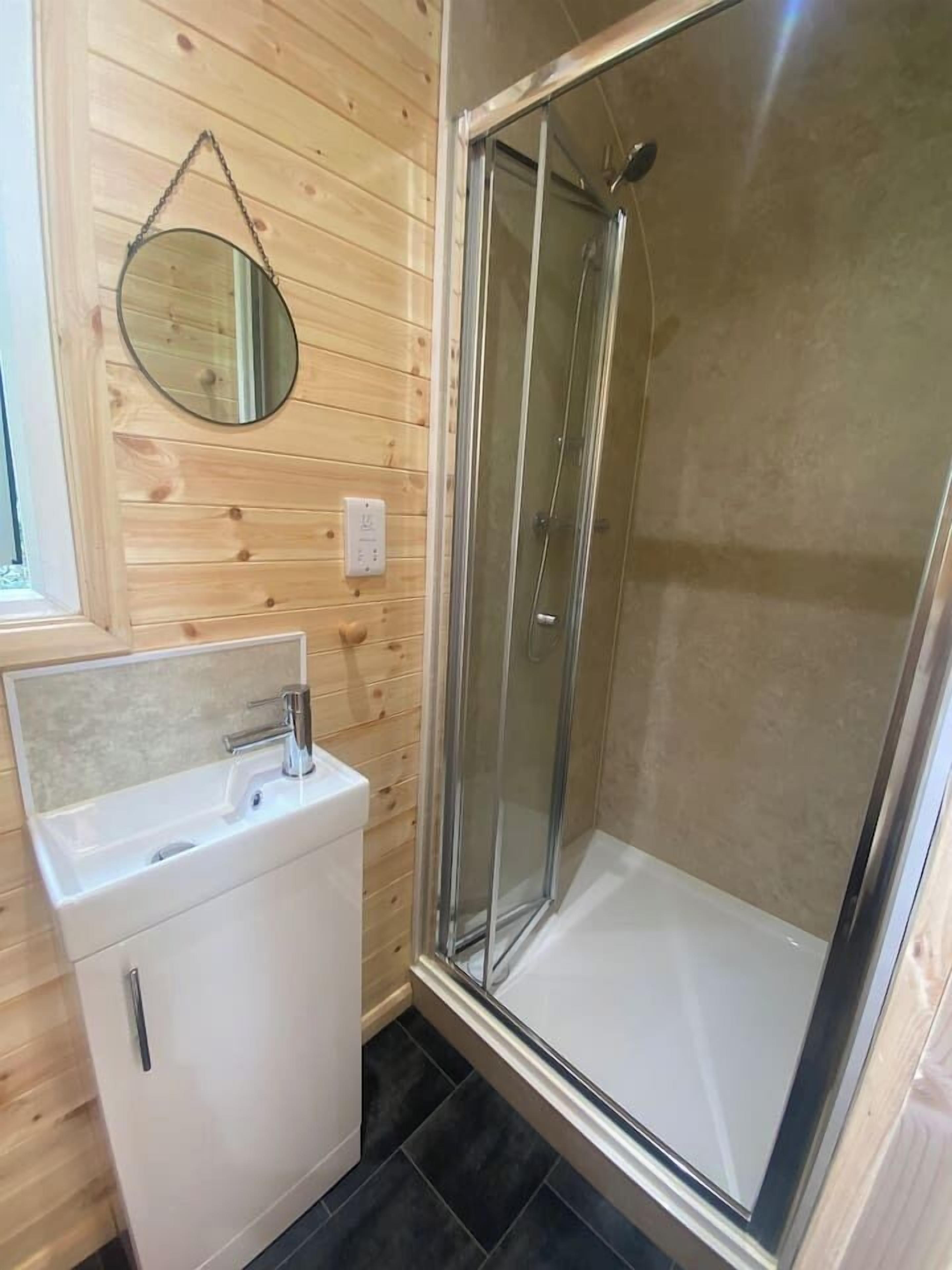 A small bathroom with a glass shower enclosure, a sink with a mirror, and wooden paneling.
