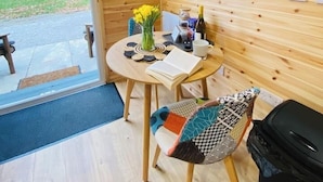 Quarto aconchegante com decoração em madeira, TV de tela plana na parede, uma mesa redonda com um livro, um vaso com flores e uma cadeira com almofada estampada.