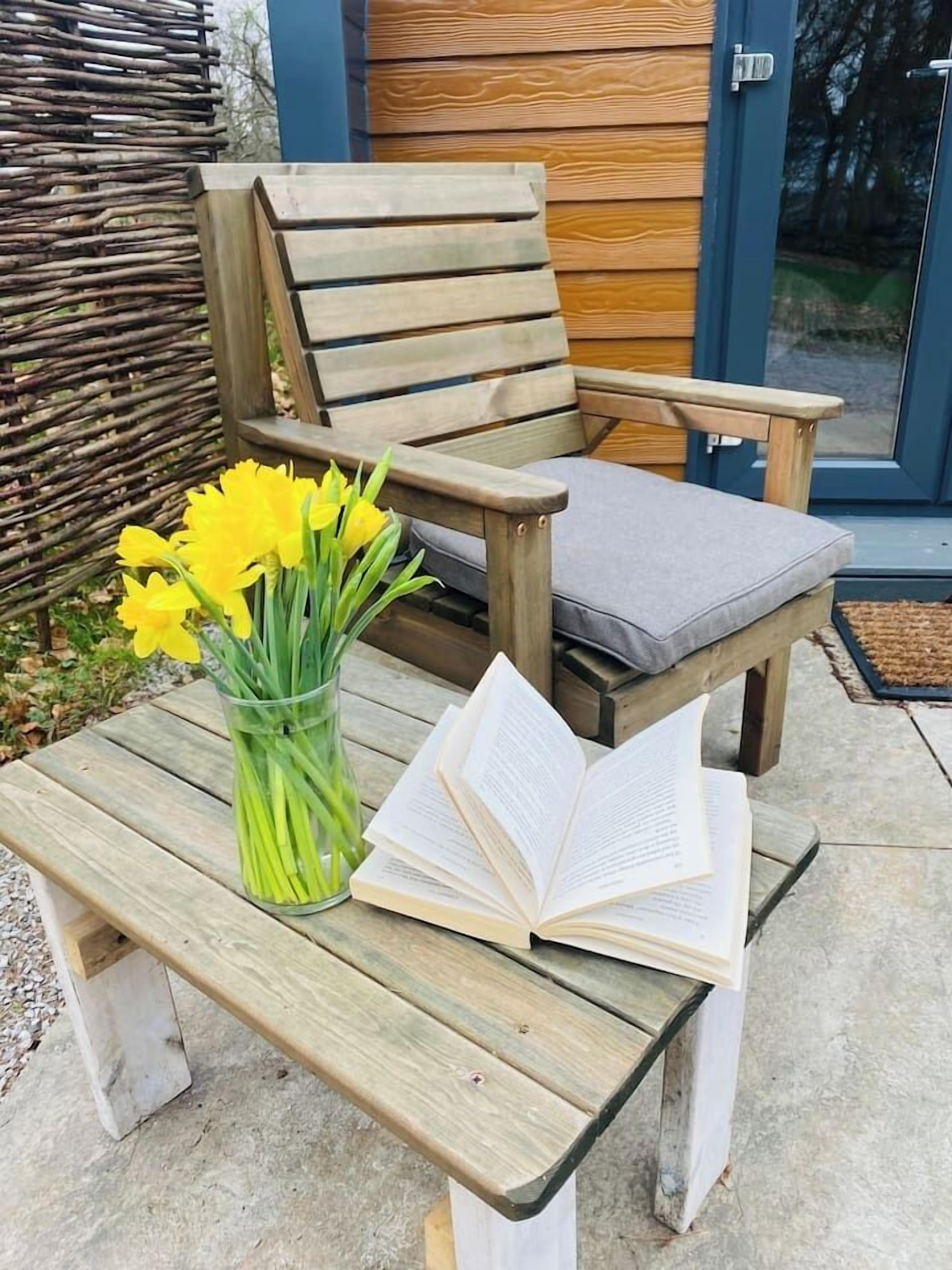 Una sedia di legno con un cuscino, un vaso di vetro con fiori gialli e un libro aperto su un tavolo di legno.