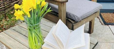 Una sedia di legno con un cuscino, un vaso di vetro con fiori gialli e un libro aperto su un tavolo di legno.