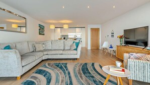 A modern living room with a sofa, a coffee table, a TV, and a kitchen in the background. - 3 Bed in Torpoint (oc-74300) (Kingsand)