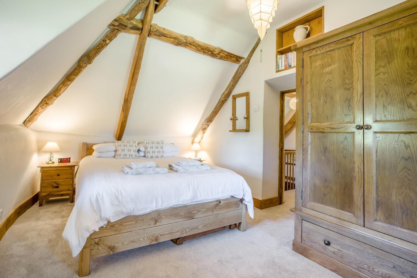 A bedroom with a wooden bed, a nightstand with a lamp, a wooden wardrobe, and a sloped ceiling with exposed beams.