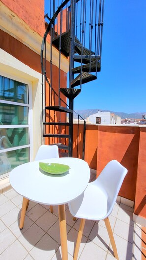 A balcony with a white table and chairs, a spiral staircase, and a view of distant buildings. - Sol Suites Loft Penthouse Apartment (Fuengirola)