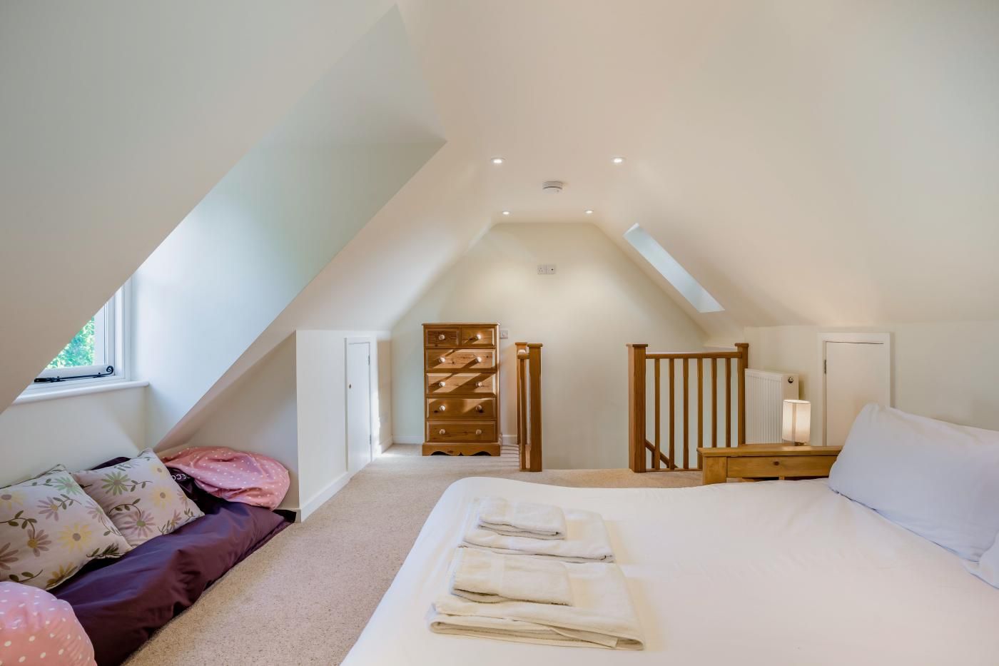 A bedroom with a sloped ceiling, a bed with white linens, a wooden dresser, a nightstand, and a window.