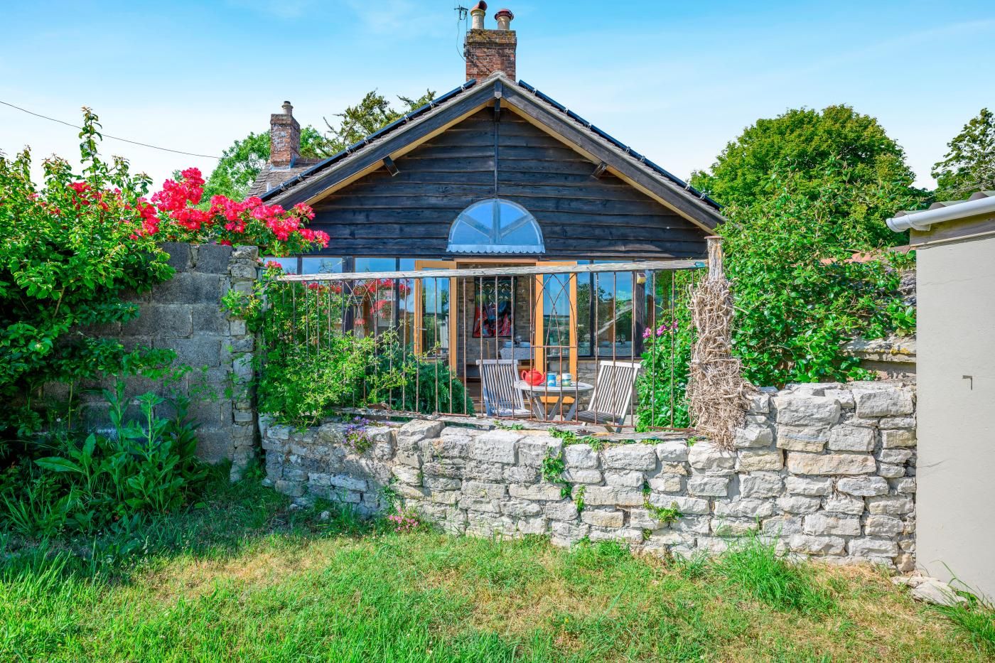 A wooden house with a glass conservatory, stone wall, and lush greenery.