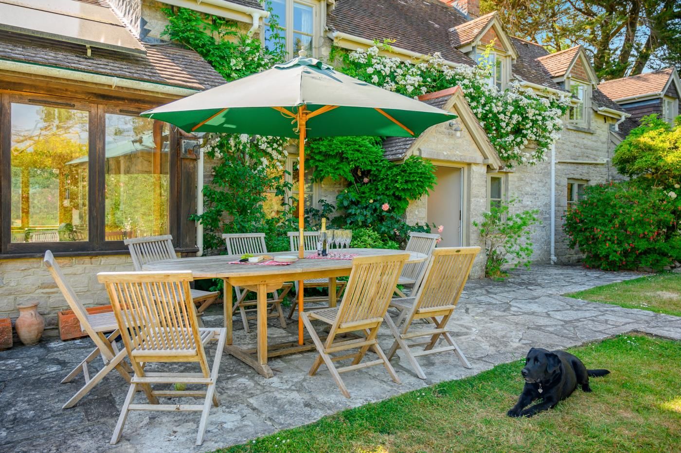 Een terras buiten met een houten tafel en stoelen, een groene parasol en een hond.