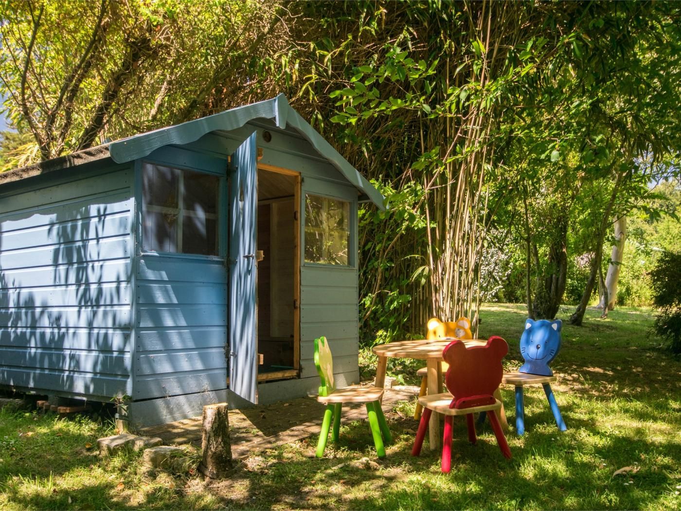 A small blue cabin with a door open, surrounded by a grassy area with a wooden table and colorful chairs.