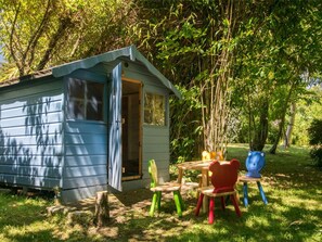A small blue cabin with a door open, surrounded by a grassy area with a wooden table and colorful chairs. - 5 Bed in Wareham (oc-dc061) (Woolgarston)