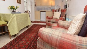 Ein gemütliches Wohnzimmer mit einer Couch, einem Esstisch mit grüner Tischdecke, einer Küche im Hintergrund und einem gemusterten Teppich.