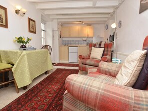 A cozy living room with a sofa, a dining table with a green tablecloth, a kitchen in the background, and a patterned rug. - 1 Bed in Bridport (oc-dc178) (Burton Bradstock)