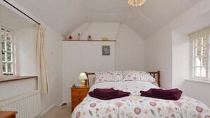 Ein Schlafzimmer mit Bett, Nachttisch, Kommode, Heizkörper, einem Fenster mit Vorhängen, einem Regal mit Dekoration und einer Deckenleuchte.