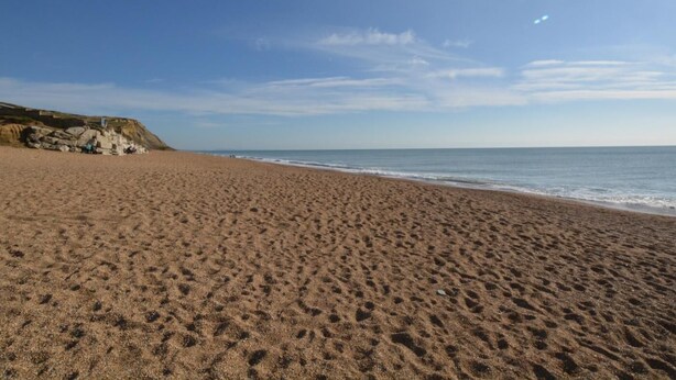 Ein weiter Sandstrand mit sanften Wellen, blauem Himmel und im Hintergrund stehenden Felsen.