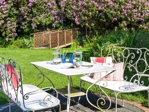 An outdoor seating area with metal chairs and a table, surrounded by lush greenery and flowering bushes. - 6 Bed in Workington (oc-sz151) (Lamplugh)