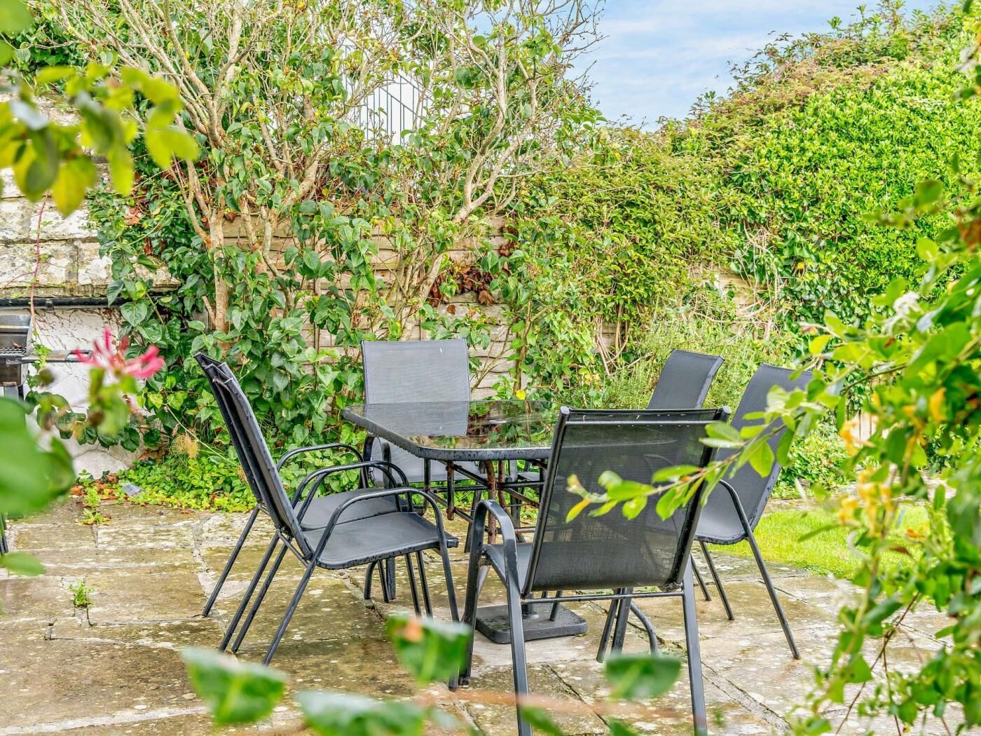 An outdoor patio with a table and four chairs, surrounded by lush greenery and a stone wall.