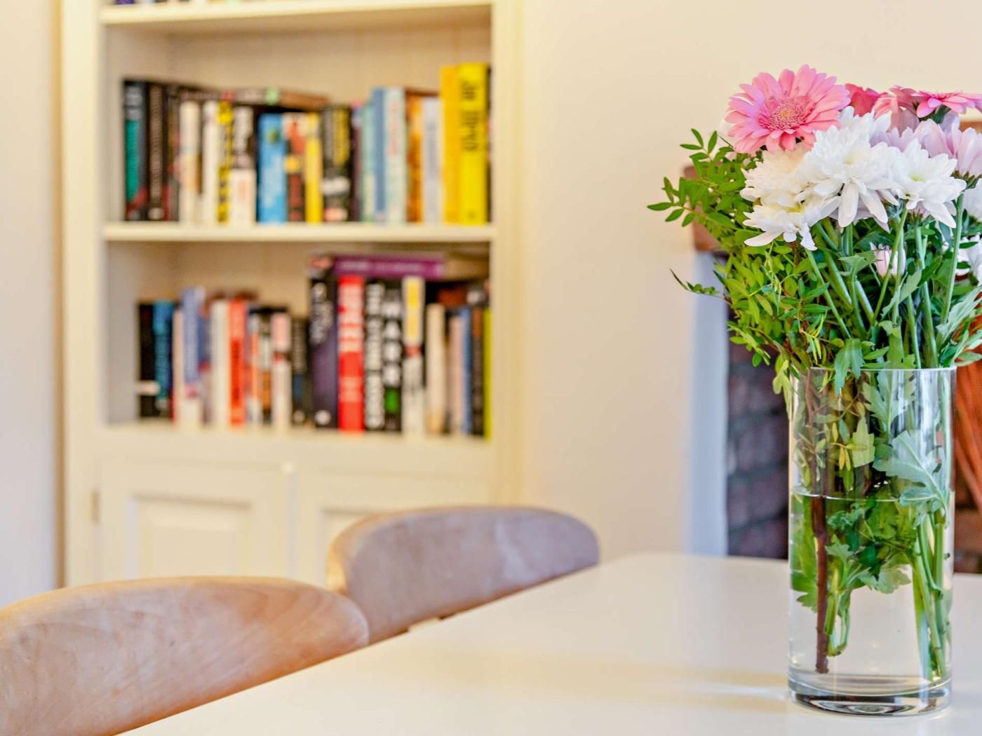 Een glazen vaas met bloemen op een tafel, een boekenkast met boeken op de achtergrond, en een effen muur.