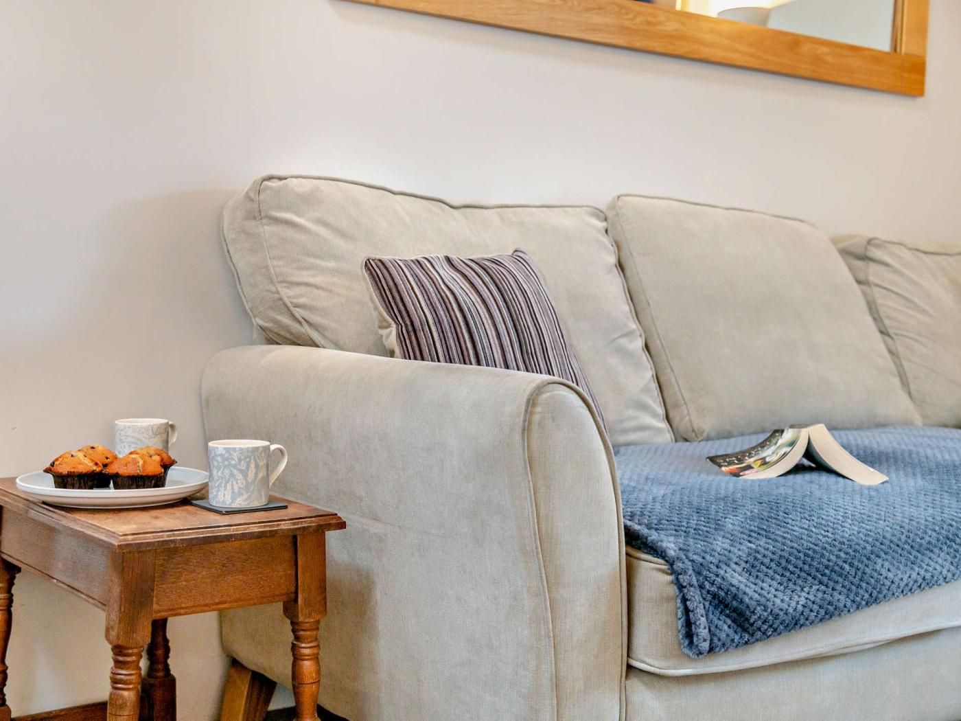 A beige sofa with a striped pillow and a blue blanket, a wooden side table with a plate of muffins and a mug, and a book.