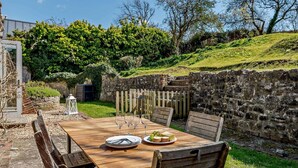 Un espacio de comedor al aire libre con una mesa de madera dispuesta para dos personas, rodeado de muros de piedra y follaje.
