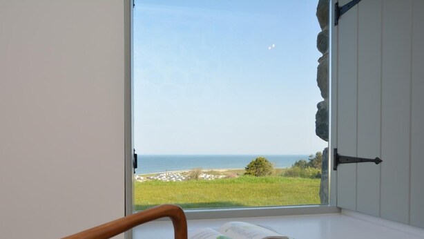 A room with a large window overlooking a coastal landscape, a wooden chair, and a book on a windowsill. - 3 Bed in Llwyngwril (oc-75025) (Llwyngwril)