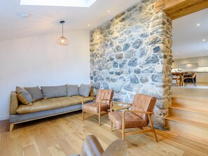 A modern living space with a stone wall, leather sofa, and wooden chairs. - 3 Bed in Llwyngwril (oc-75025) (Llwyngwril)