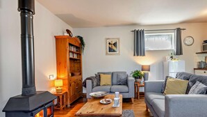 A cozy living room with a wood-burning stove, a bookshelf, a grey sofa, a wooden coffee table, and a kitchen area with a refrigerator and microwave. - 1 Bed in Dorchester (oc-75157) (Winfrith Newburgh)