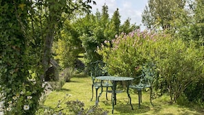 Um jardim com mesa e cadeiras de metal, rodeado por diversas plantas e flores.