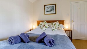 Un letto rifatto con cura, con una coperta blu, cuscini floreali e asciugamani arrotolati su una testiera in legno.