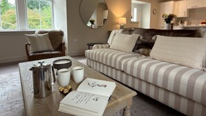 Ein gemütliches Wohnzimmer mit einem gestreiften Sofa, einem Couchtisch mit Büchern und einem Wasserkocher, einem Spiegel und Blick in die Küche.