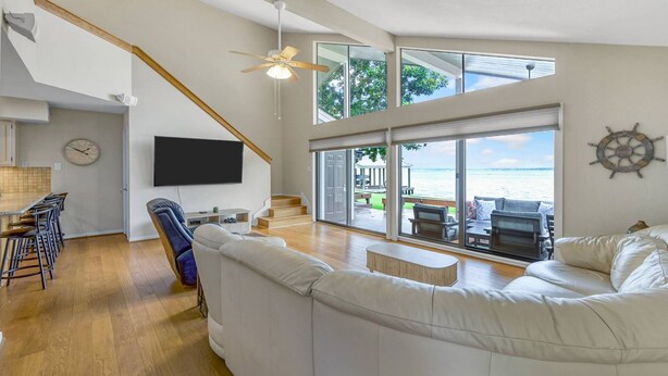 A spacious living area with a staircase, a flat-screen TV, a large white sofa, and a view of the sea through large windows. - Lakefront Townhome | Private Boat Slip | Swim + Fish | Pool Access | Near Ren Fest | Sleeps 8 (Montgomery)