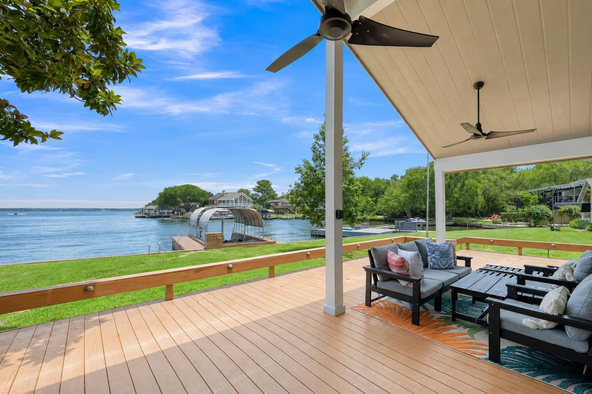 Eine überdachte Terrasse mit Gartenmöbeln, einem Deckenventilator und Blick auf einen See mit Booten.